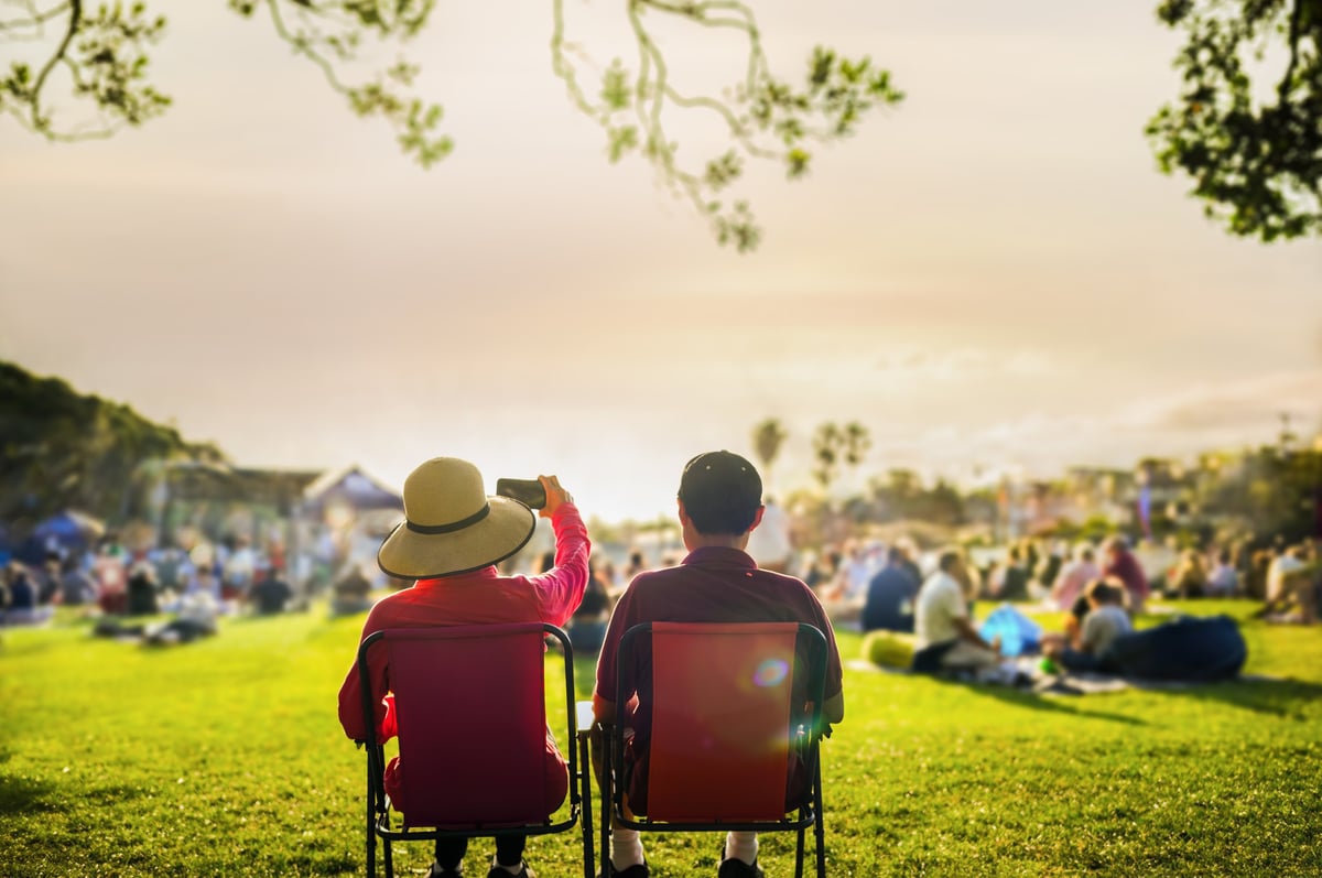 Couple taking photos using smartphone at sunset, unrecognizable crowd of people listen to the music concert in the park.