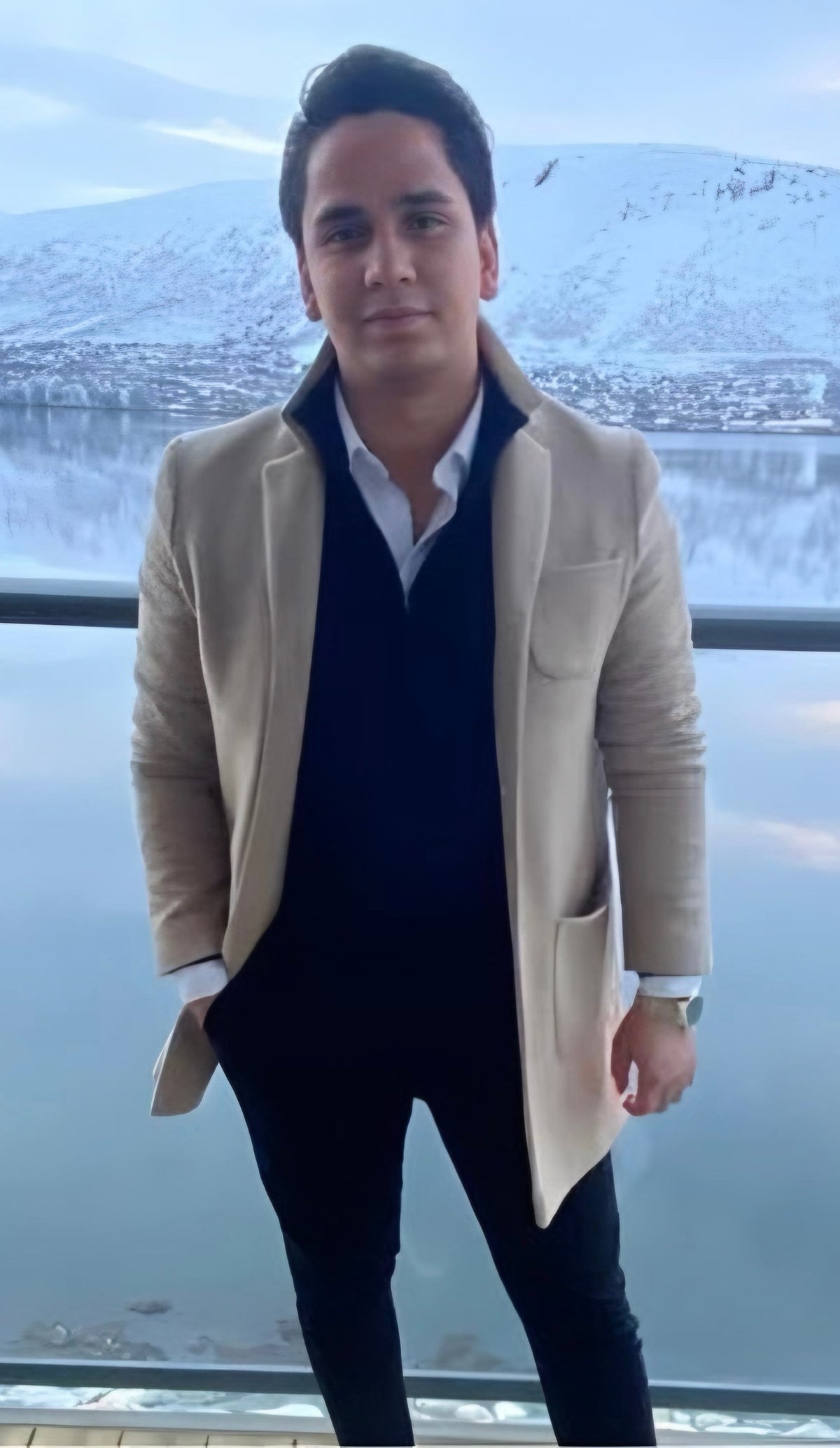 Man wearing beige cardigan and dark shirt standing in front of snowy mountain and river landscape